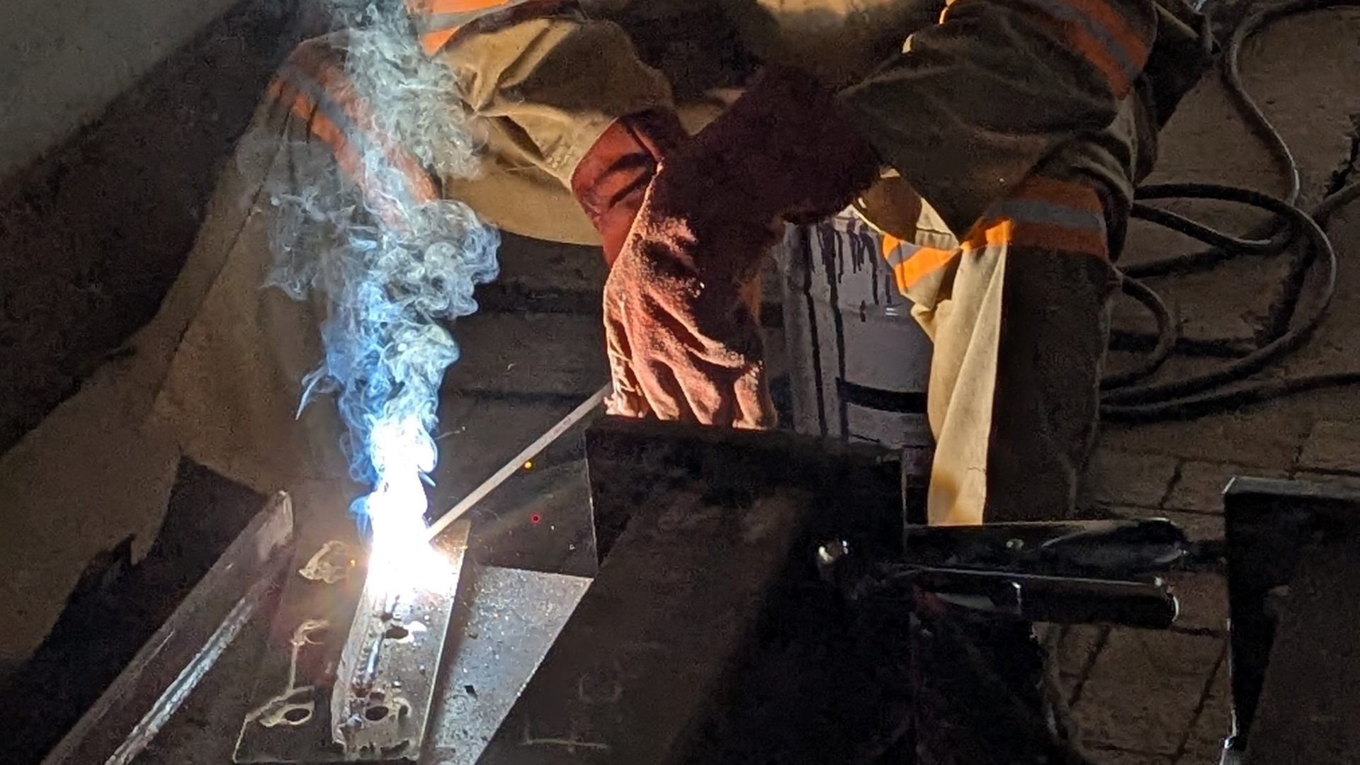 Welder in full protective gear welding a structural steel beam assembly, bright arc and rising smoke, face fully covered by welding helmet