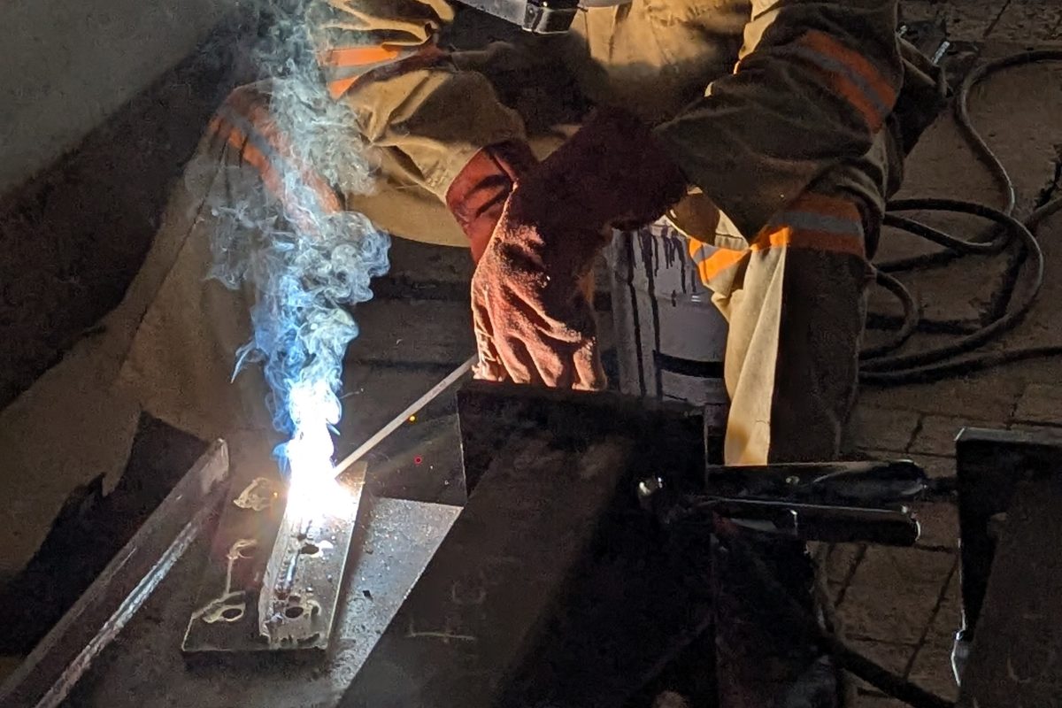 Welder in full protective gear welding a structural steel beam assembly, bright arc and rising smoke, face fully covered by welding helmet